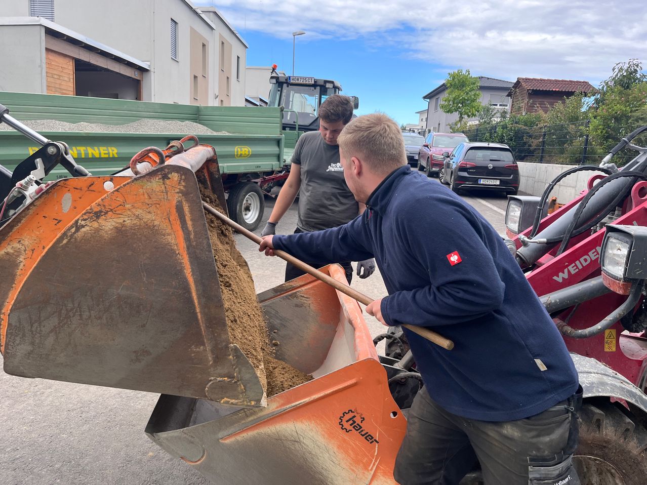 Zwei Männer bedienen einen Ladeschaufel, einer hält einen Holzstab und der andere beobachtet. Dahinter sind ein grüner LKW und ein rotes Maschinenfahrzeug geparkt. Autos sind am Straßenrand geparkt.