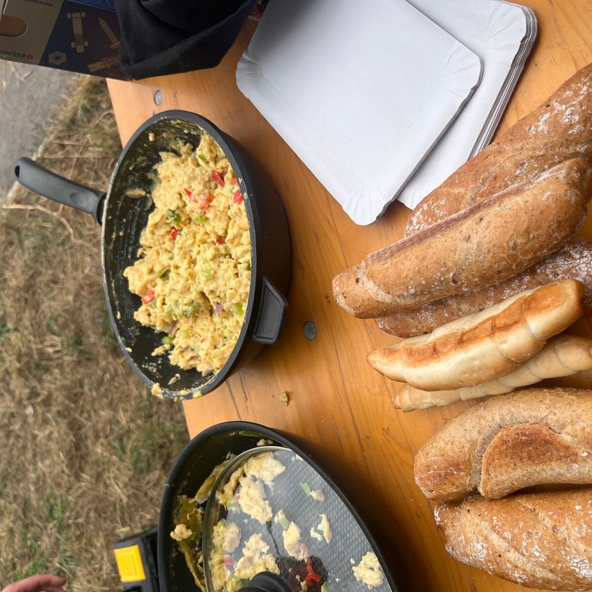 Ein Holztisch mit einer Bratpfanne mit Rührei und einem Stapel Baguettes auf der rechten Seite. Zwei weiße Servietten liegen auf dem Tisch.