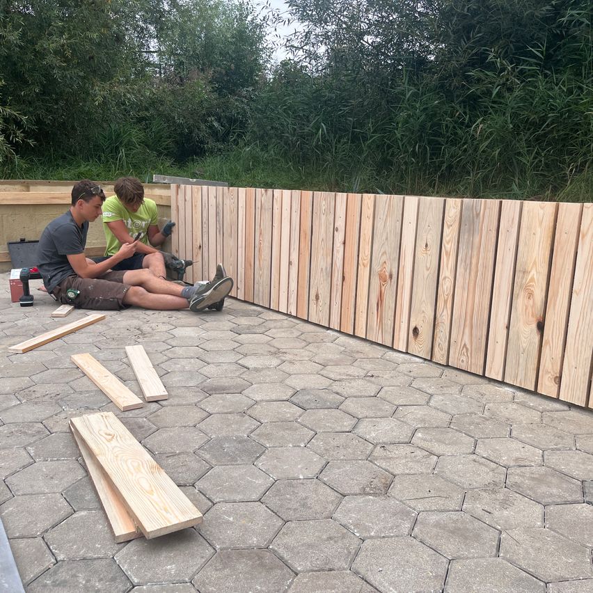 Zwei Männer sitzen auf dem Boden und arbeiten an einem Holzzaun, mit Holzstücken und Werkzeugen um sie herum, vor einer Kulisse von Grün.