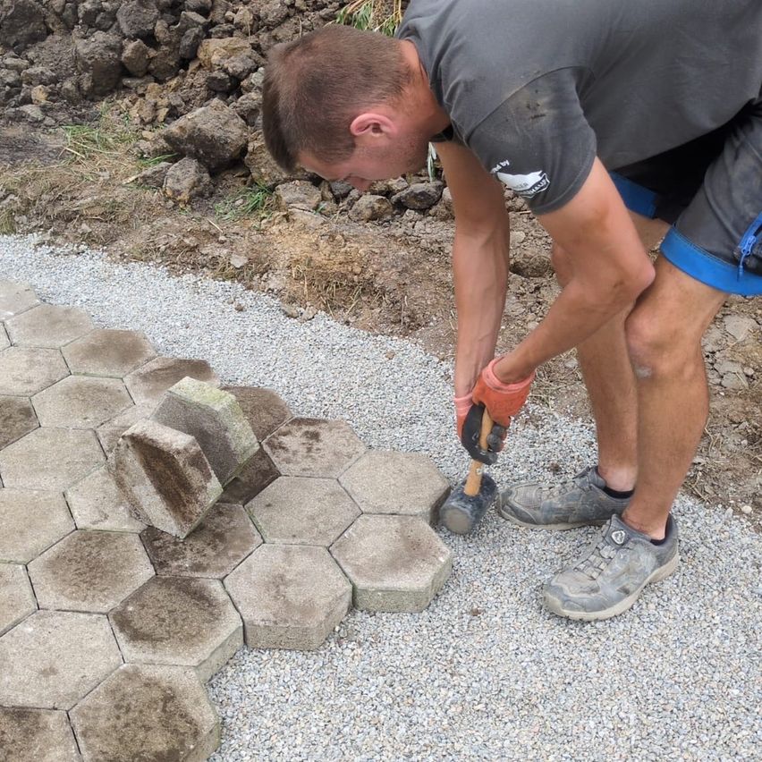 Ein Mann installiert einen hexagonalen Betonweg und benutzt dabei einen Hammer. Er trägt Handschuhe und Turnschuhe.