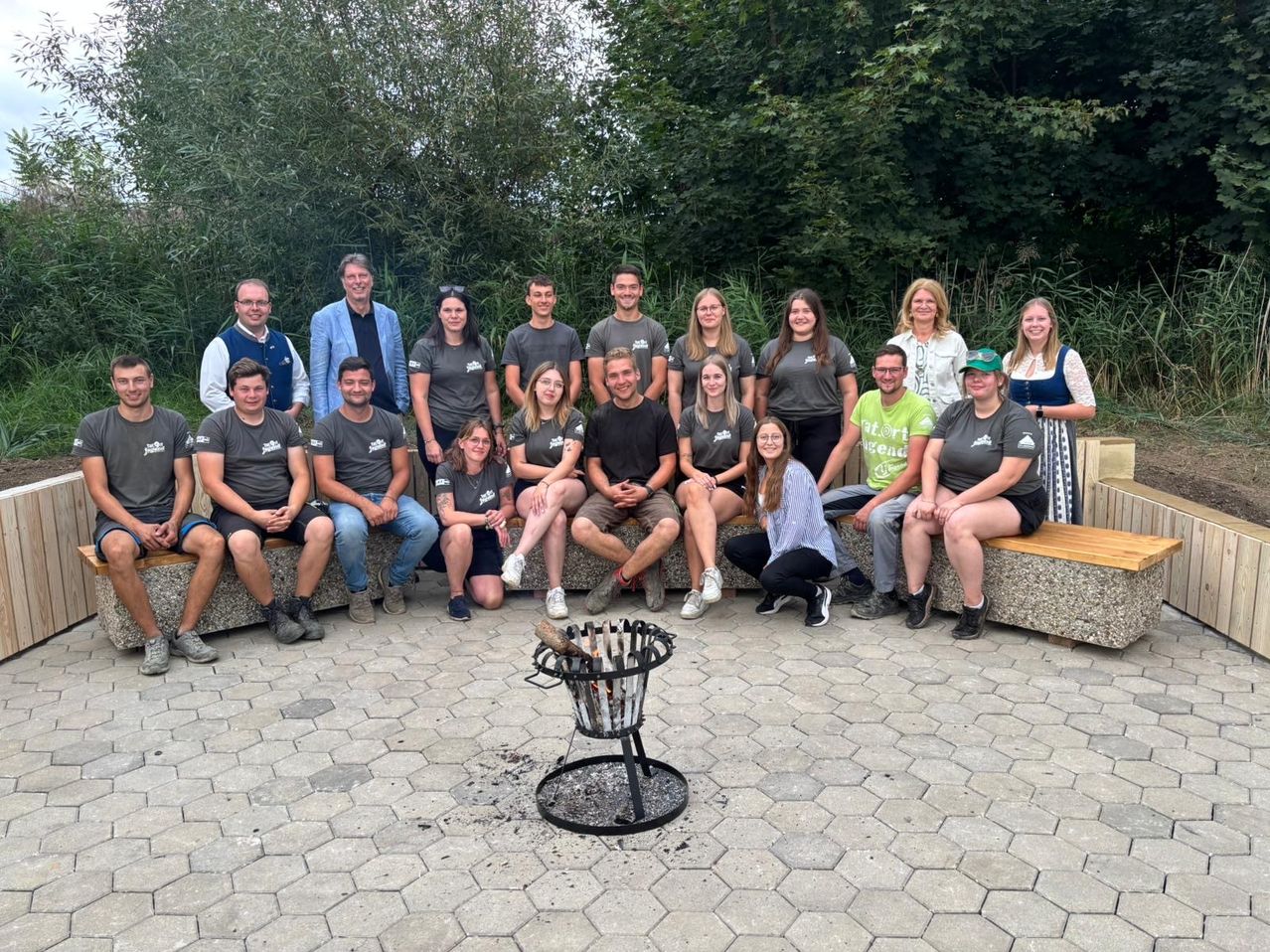 Eine Gruppe von Menschen in gleichen Shirts posiert für ein Foto neben einem Feuerkorb, umgeben von Grün.
