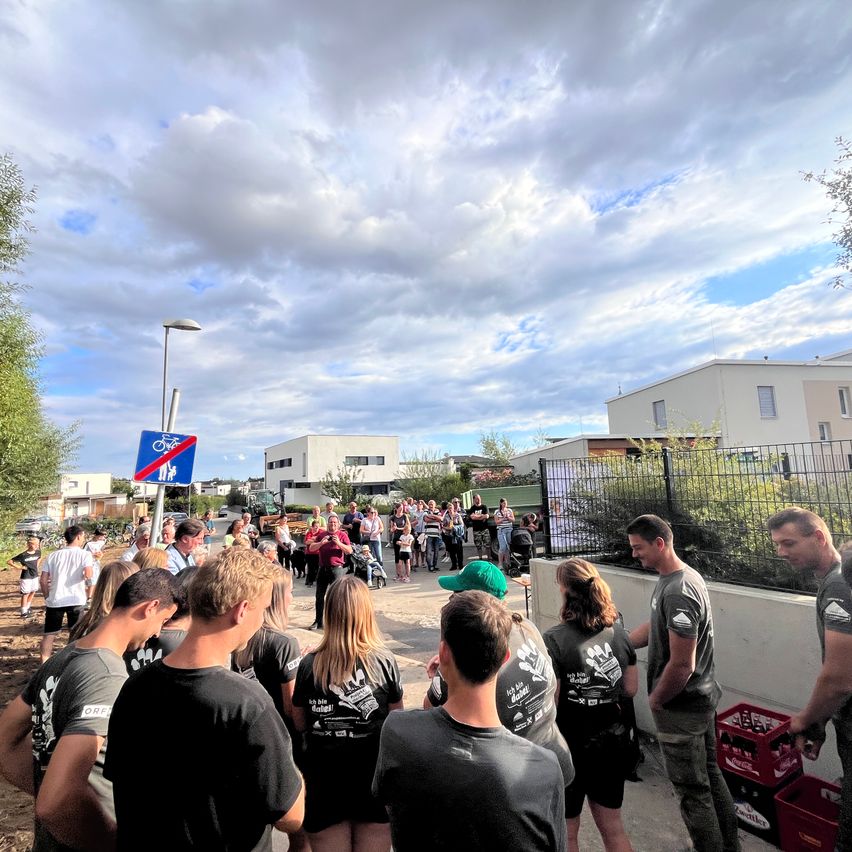 Eine Gruppe von Menschen in gleichen T-Shirts steht draußen in der Nähe einer Mauer. Sie schauen einen Mann in einer grünen Mütze an, der zu ihnen spricht. Dahinter stehen weitere Menschen und Gebäude. Ein Fahrradschild steht links an einem Pfosten.