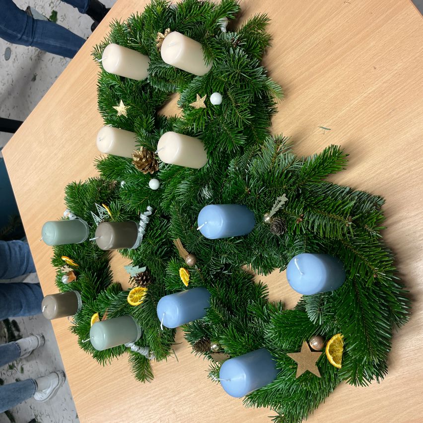 Ein Kranz aus immergrünen Pflanzen, blauen und weißen Kerzen sowie dekorativen Sternen und Orangen auf einem Holztisch.