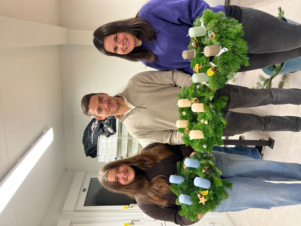 Drei Personen lächeln und posieren für ein Foto. Sie halten eine Girlande mit Kerzen und Tannenzapfen. Sie befinden sich in einem Raum mit weißen Wänden und einer weißen Decke. Hinter ihnen steht eine schwarze Jacke und ein weißer Regal.