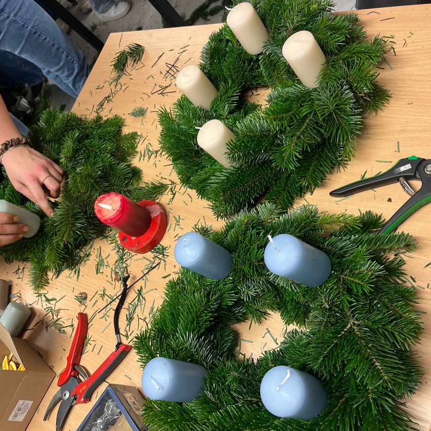 Ein Holztisch zeigt mehrere Kränze aus grünen Tannennadeln und weißen und blauen Kerzen. Ein Kranz hat eine rote Kerze in der Mitte. Werkzeuge wie Scheren und Zangen sind herumliegen.