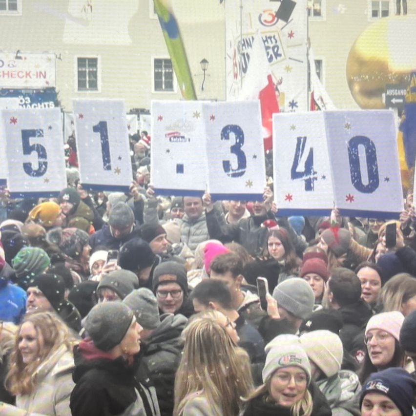 Eine Menschenmenge im Freien, die Schilder mit den Zahlen 5, 1, 3, 4 und 0 hochhält. Einige tragen Winterkleidung, andere halten Telefone. Im Hintergrund ist ein Gebäude mit einem Schild zu sehen, auf dem Check-In steht.