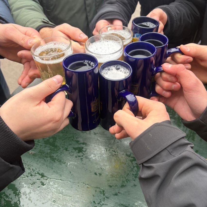 Eine Gruppe von Menschen versammelt sich um einen Tisch, jeder hält eine blaue Tasse mit Bier, vermutlich bei einer Feier oder einem gesellschaftlichen Treffen.