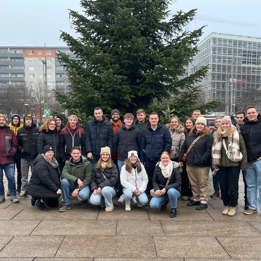 Eine Gruppe von Menschen posiert für ein Foto vor einem Weihnachtsbaum. Sie tragen alle Winterkleidung. Der Weihnachtsbaum ist geschmückt. Hinter ihnen stehen Gebäude.