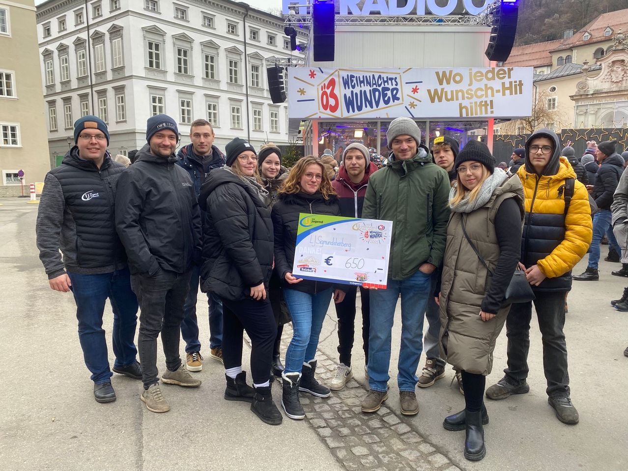 Eine Gruppe von Menschen posiert für ein Foto vor einem Banner, das 'Weihnachtswunder' besagt. Eine Frau hält ein Schild mit einem Scheck über 650 Euro. Sie sind warm gekleidet und stehen auf Kopfsteinpflaster. Dahinter steht ein Gebäude mit vielen Fenstern.