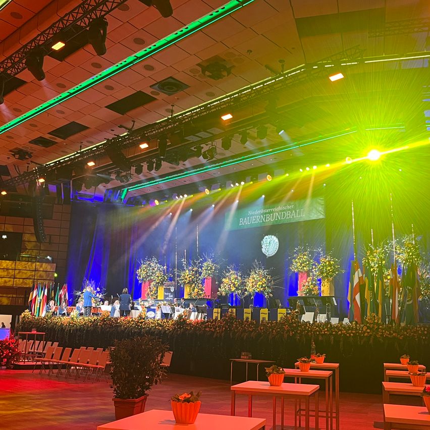 A stage with a banner reading 'Niedernsterreidischer Bauernbundball'. An orchestra is performing on stage with a spotlight. Many flags are behind the stage. The stage is decorated with flowers. There are rows of chairs on the left side.