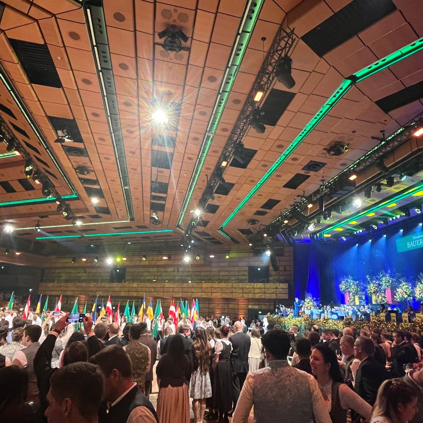 A crowd of people gathered in a large hall, holding flags and taking photos. The stage has a green backdrop with blue lights and a sign reading 'BAUER'. The ceiling has a series of spotlights.
