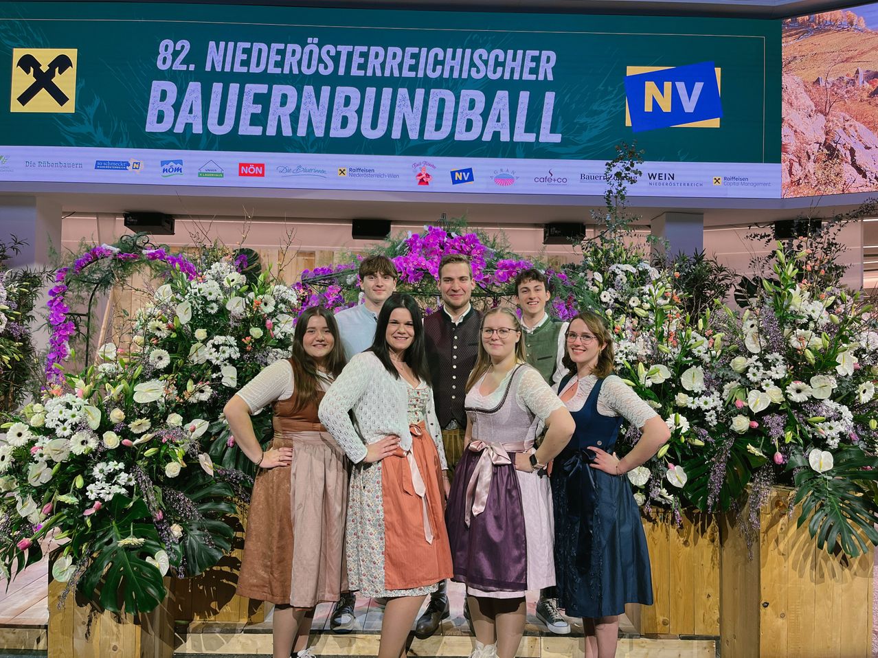 Group photo of six people in traditional Bavarian attire standing in front of a floral backdrop with a sign reading '82. Niederosterreichischer Bauernbundball.'