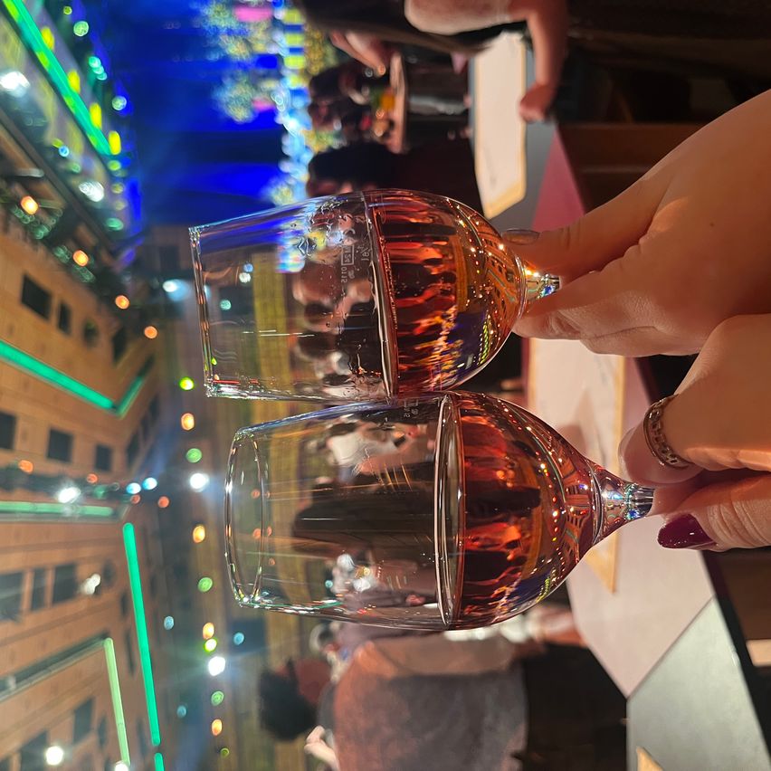 Two hands holding wine glasses filled with rose wine, reflecting colorful lights from the ceiling, with blurred people in the background.
