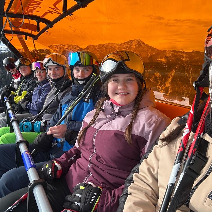Eine Gruppe von Skifahrern sitzt zusammen auf einem überdachten Fahrzeug, alle tragen Helme und Schutzbrillen. Sie halten Skistöcke und scheinen zu lächeln. Das Fahrzeug ist orange mit einem Metallrahmen. Hinter ihnen sind Berge unter einem warmen, orangefarbenen Himmel sichtbar.