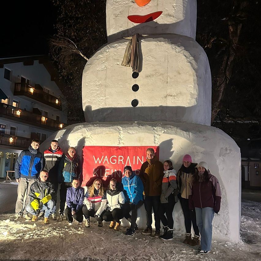 Eine Gruppe von Menschen in Winterkleidung posiert für ein Foto vor einem großen Schneemann, mit einem Banner hinter ihnen, das 'Wagrain Klein' steht.