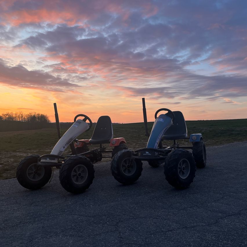 Two go-karts are parked on a road with a sunset in the background. Both vehicles have black seats and wheels.