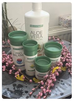 Mehrere weiße und grüne Flaschen mit verschiedenen Etiketten sind auf einem Tisch angeordnet, umgeben von rosa Blumen. Eine der Flaschen hat ein Etikett, das Aloe Vera Trinkgel sagt.