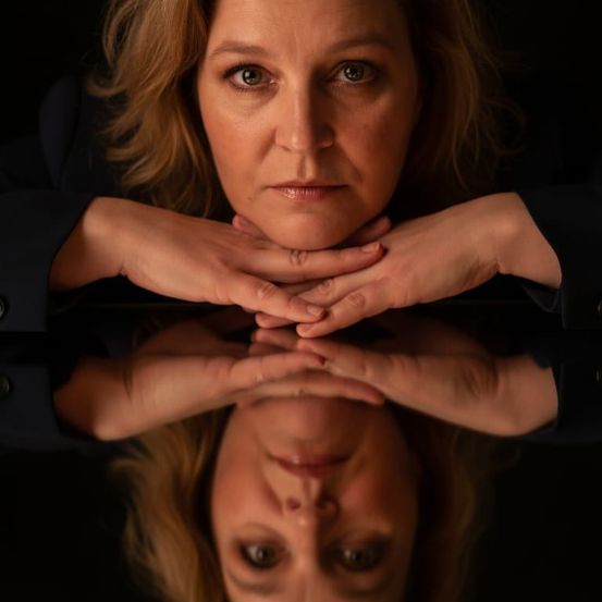 Eine Frau mit blonden Haaren posiert für ein Foto mit den Händen am Kinn. Sie hat einen ernsten Gesichtsausdruck und trägt ein schwarzes Outfit. Ihr Spiegelbild ist auf der Glasoberfläche sichtbar.