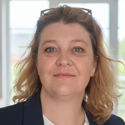 Eine Frau mit blondem Haar steht in einem Raum mit einem Bücherregal und Fenstern. Sie trägt einen marineblauen Blazer über einem weißen Blusenshirt und hat eine Brille auf dem Kopf.