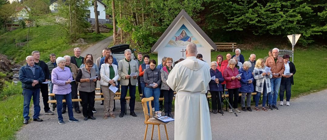 Ein Priester steht vor einer Kapelle mit einer Menschenmenge, alle halten Papiere, bei einer Zeremonie im Freien.