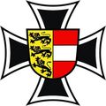 Das Bild zeigt ein heraldisches Emblem mit einem Andreaskreuz und einem zentralen Schild. Der Schild hat einen roten und weißen Streifen unten und ein gelbes Feld mit einem schwarzen Löwen darüber.
