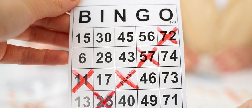 Eine Person hält einen Bingo-Karte mit den Zahlen 15-73, einige Zahlen sind mit roten X's markiert. Im Hintergrund sitzen ein Mann und eine Frau am Tisch.