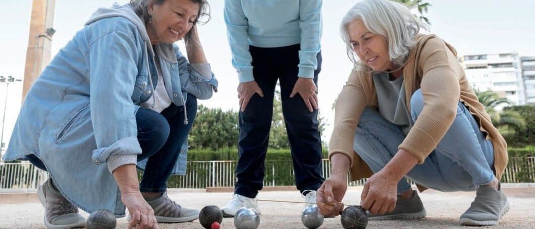 Drei ältere Frauen spielen Boule auf einem sandigen Platz, eine kniet und zwei stehen. Sie sind von Boules umgeben und im Hintergrund befindet sich ein Holzzaun.