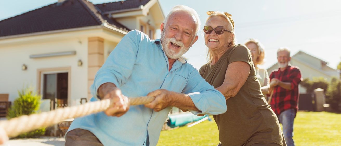 Älteres Paar zieht an einem Seil und lächelt auf einem Rasen vor einem Haus.