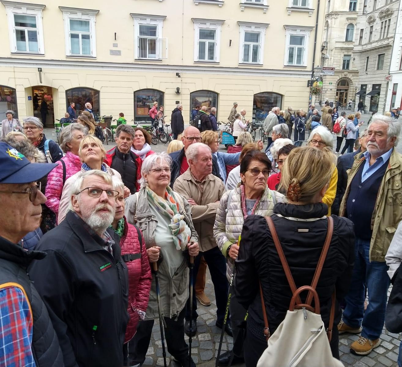 Eine Gruppe von Menschen ist an einem Außenort versammelt, einige tragen Brillen und haben Rucksäcke dabei. Sie stehen vor einem Gebäude, vor dem Fahrräder geparkt sind.