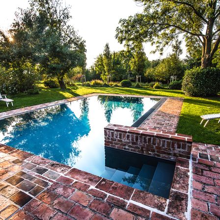 Ein rechteckiger Swimmingpool aus Ziegelsteinen ist von üppiger Grünfläche umgeben, mit einer kleinen Vertiefung in der Mitte, die zu einer Treppe führt. Bäume und Pflanzen sind im Hintergrund sichtbar.