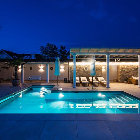 Ein moderner Außenpool mit beleuchteten Stufen und Liegestühlen, umgeben von einem Pavillon mit Steinwänden und Sonnenschirmen. Bäume und ein Haus sind im Hintergrund zu sehen.