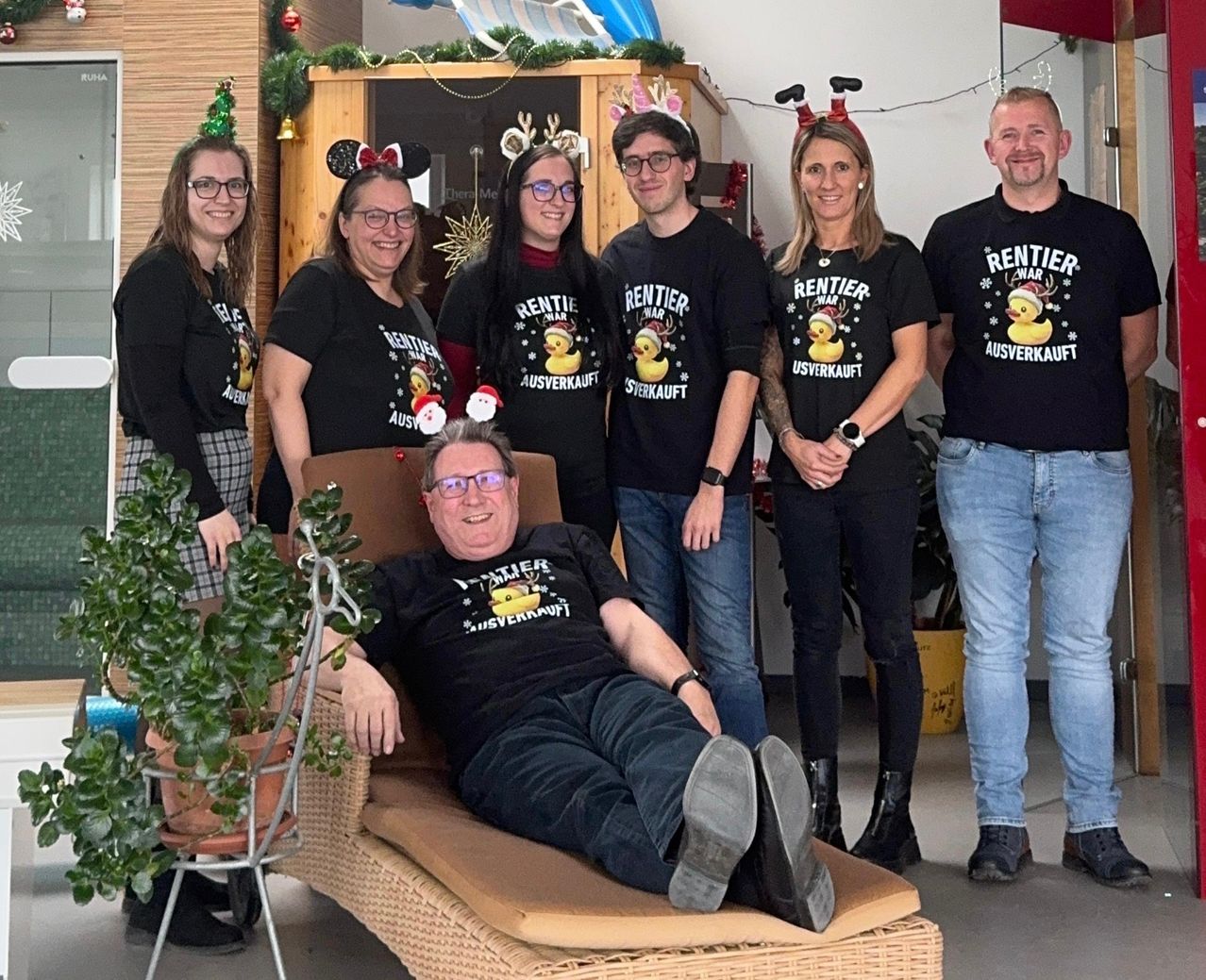 Eine Gruppe von Menschen in passenden Hemden mit Entendesign posiert für ein Foto. Sie stehen und sitzen um einen Stuhl herum. Ein Mann sitzt mit erhobenen Beinen auf einem Stuhl. Sie tragen alle eine Brille und einige tragen Stirnbänder. Hinter ihnen steht ein Weihnachtsbaum und ein hölzerner Schrank.