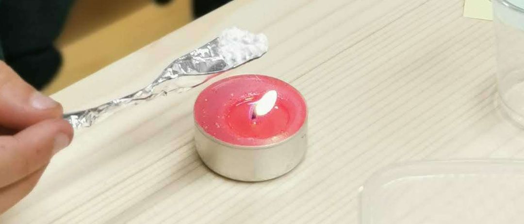 Zwei Jungen sitzen an einem Tisch und sind jeweils mit einer konzentrierten Aktivität beschäftigt. Der Junge auf der linken Seite hält ein kleines Objekt, möglicherweise eine Kerze, mit einer Pinzette. Der Junge auf der rechten Seite beobachtet etwas auf dem Tisch. Andere Kinder und Erwachsene sind im Hintergrund ebenfalls mit Aktivitäten beschäftigt.