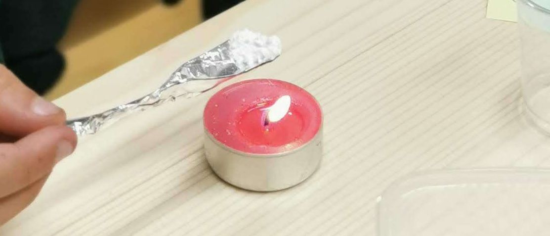 Zwei Jungen sitzen an einem Tisch und sind jeweils mit einer konzentrierten Aktivität beschäftigt. Der Junge auf der linken Seite hält ein kleines Objekt, möglicherweise eine Kerze, mit einer Pinzette. Der Junge auf der rechten Seite beobachtet etwas auf dem Tisch. Andere Kinder und Erwachsene sind im Hintergrund ebenfalls mit Aktivitäten beschäftigt.