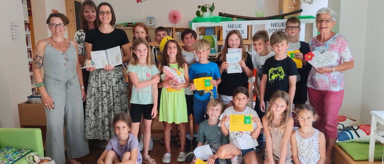 Eine Gruppe von Kindern und Erwachsenen posiert mit Urkunden in einem Klassenzimmer. Der Raum hat einen Holzboden und eine weiße Wand mit bunten Dekorationen.