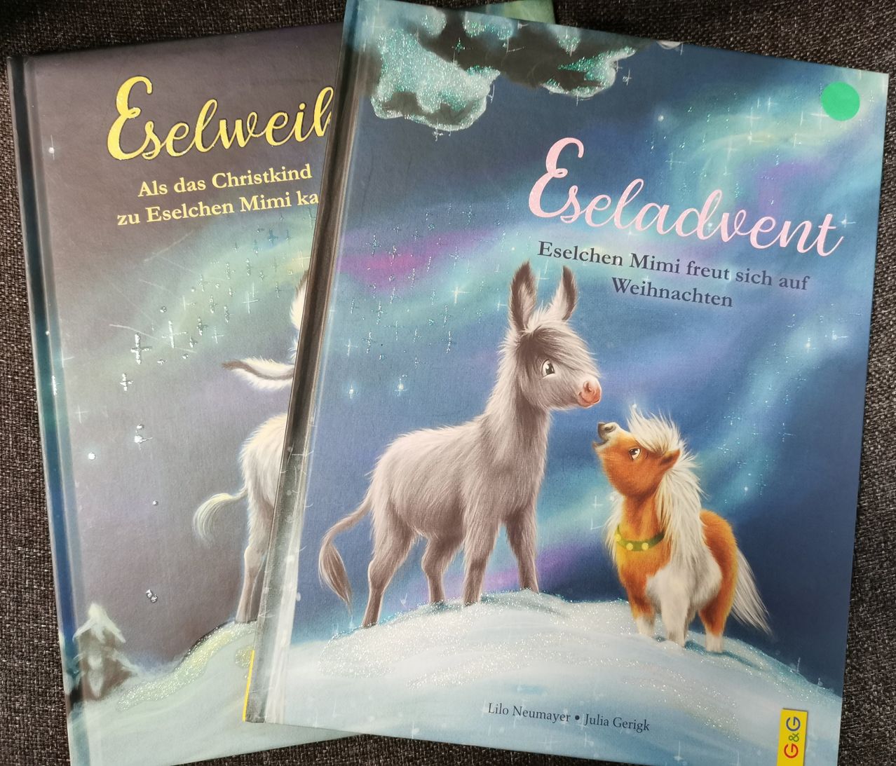 Zwei Bücher liegen auf einer braunen Oberfläche. Das Buch auf der linken Seite hat den Titel Eselweih und zeigt ein weißes Esel und ein weißes Pferd. Das Buch auf der rechten Seite hat den Titel Eseladvent und zeigt ein weißes Esel, ein braun-weißes Pony und eine verschneite Landschaft.