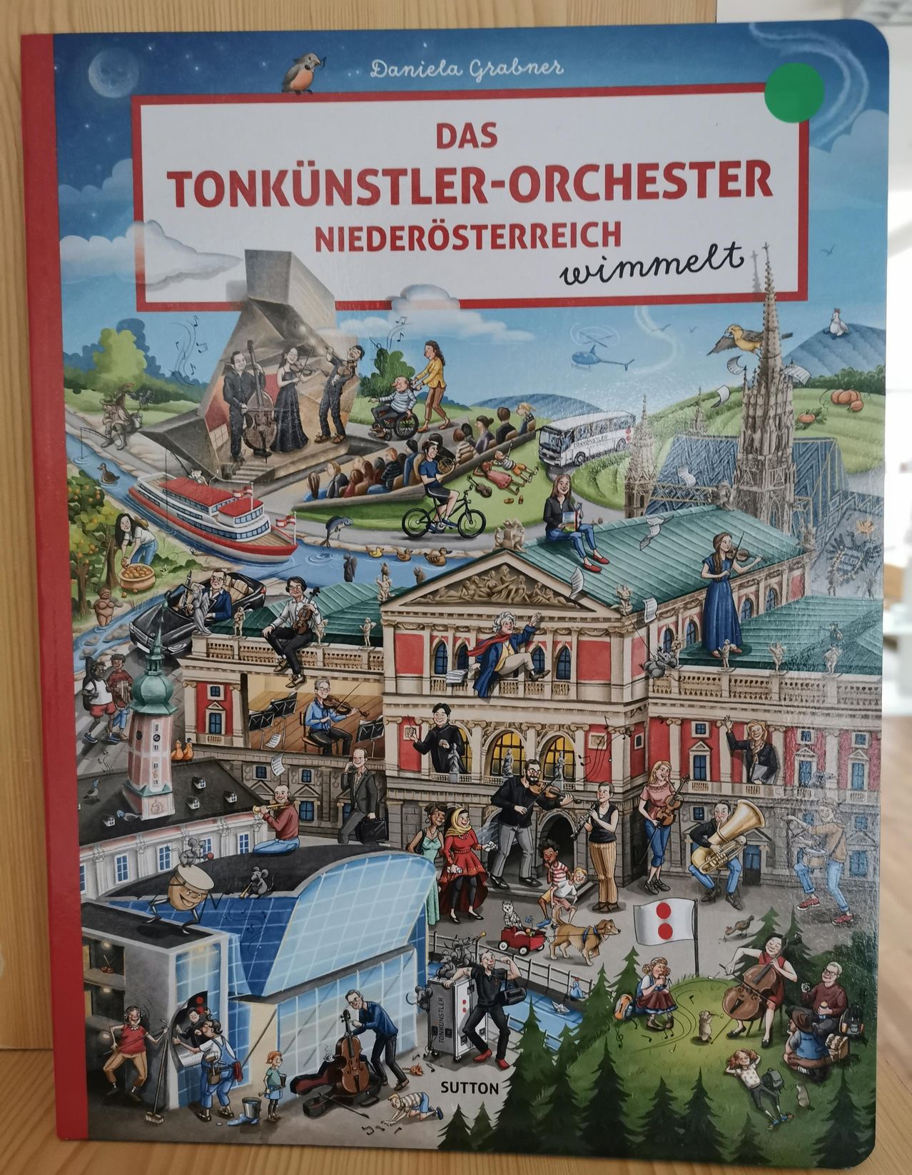 Eine bunte Illustration von Musikern an verschiedenen Orten, einschließlich eines Schlosses, eines Bootes und einer Stadt. Der Titel lautet 'Das Tonkünstler-Orchester Niederösterreic wimmelt'.