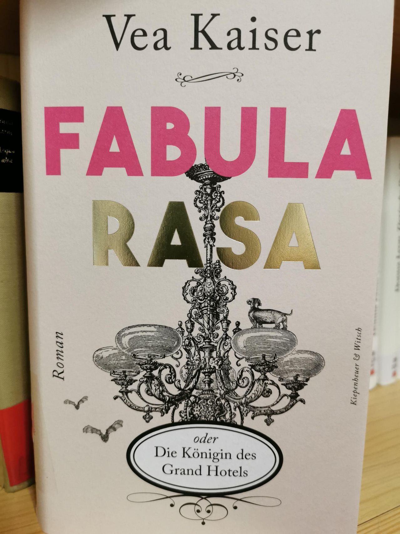 Ein Buchcover mit dem Titel 'Fabula Rasa' zeigt eine goldene Kronleuchter mit einem Hund darauf, umgeben von Fledermäusen. Es ist in pink und gold geschrieben.