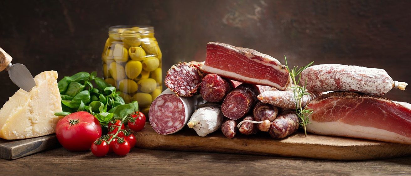 Eine Auswahl an Wurstwaren, darunter Salami und Prosciutto, liegt auf einem Holzbrett neben Tomaten und Basilikum. Dahinter ist ein Glas mit Oliven zu sehen.
