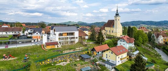 An aerial view of a small town with houses and a church under a cloudy sky. The houses have various colors, and a garden is visible in front of a white building.