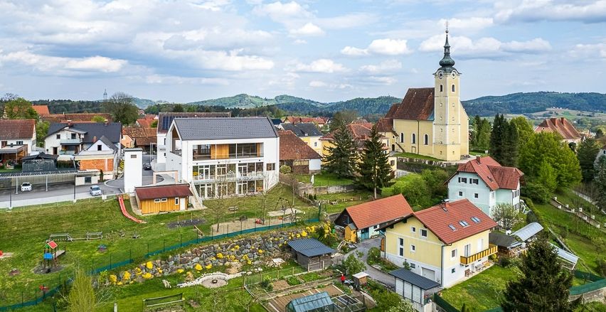Eine Luftaufnahme einer Kleinstadt mit Häusern und einer Kirche unter einem bewölkten Himmel. Die Häuser haben verschiedene Farben, und ein Garten ist vor einem weißen Gebäude zu sehen.