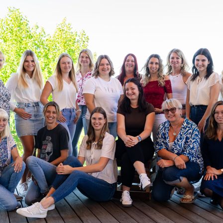 Eine Gruppe von Frauen in lässiger Kleidung posiert für ein Foto auf einer Holzterrasse unter Bäumen. Einige sitzen, andere stehen.