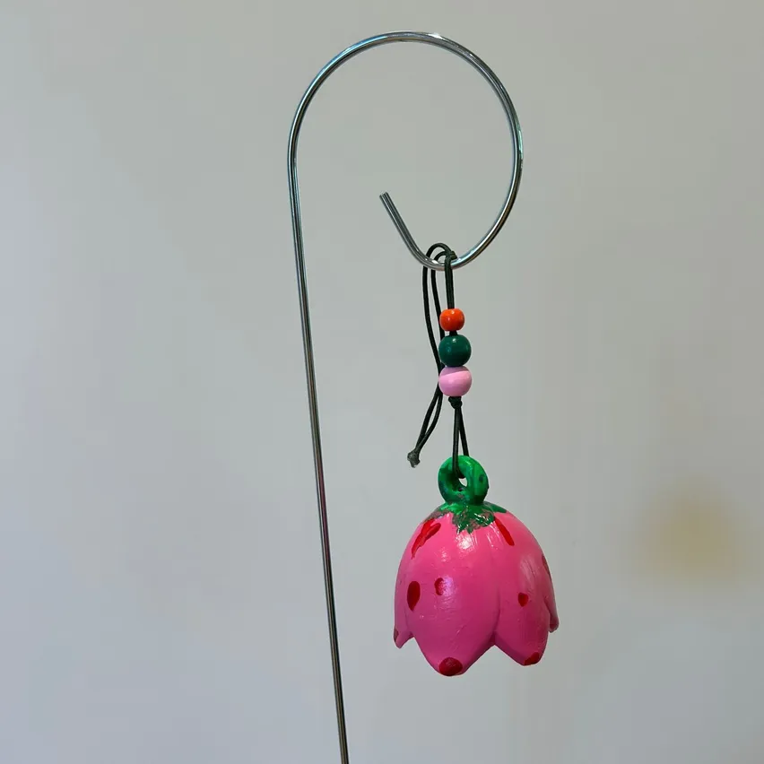Ein rosa, gepunkteter blumenförmiger Anhänger hängt an einem silbernen Ring an einem Stab vor weißem Hintergrund. Der Blume sind bunte Perlen angebracht.