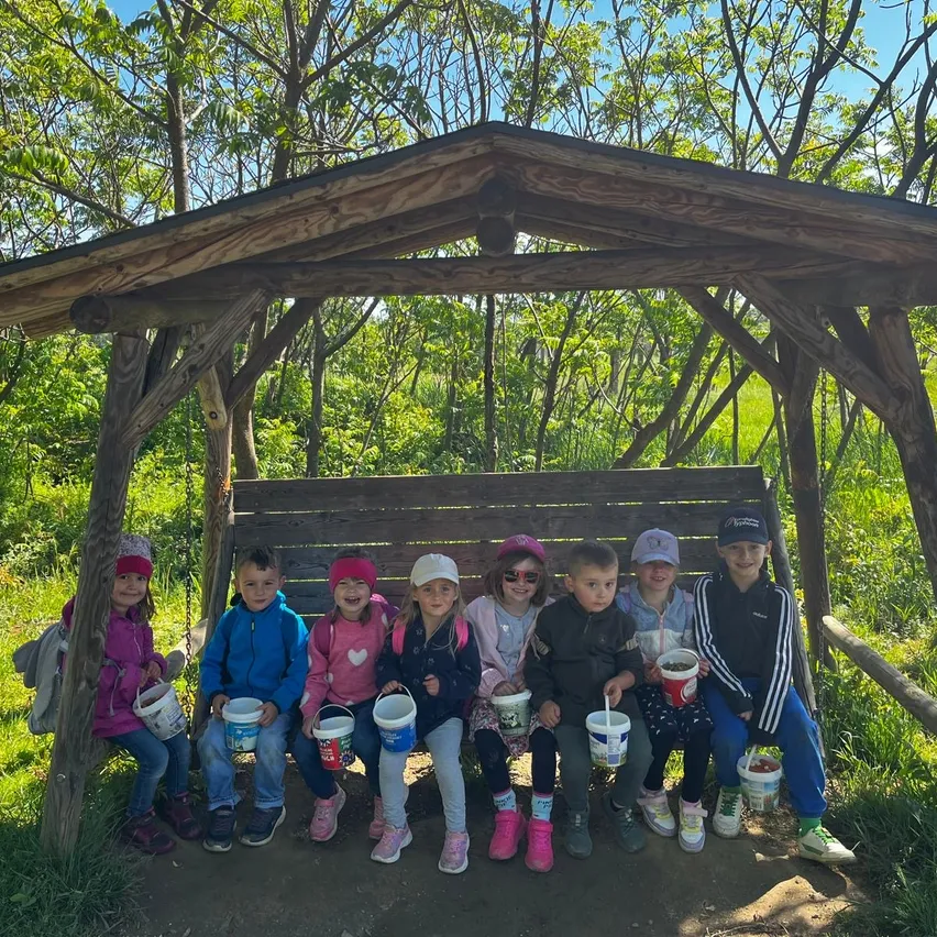 Eine Gruppe von Kindern in übereinstimmenden Jacken sitzt auf einer Holzbank in einem Wald. Sie halten Eimer, möglicherweise gefüllt mit etwas. Die Bank ist von einem Holzdach bedeckt.