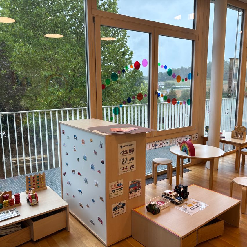 A playroom with wooden furniture, a large whiteboard with stickers, and a view of a tree and balcony.