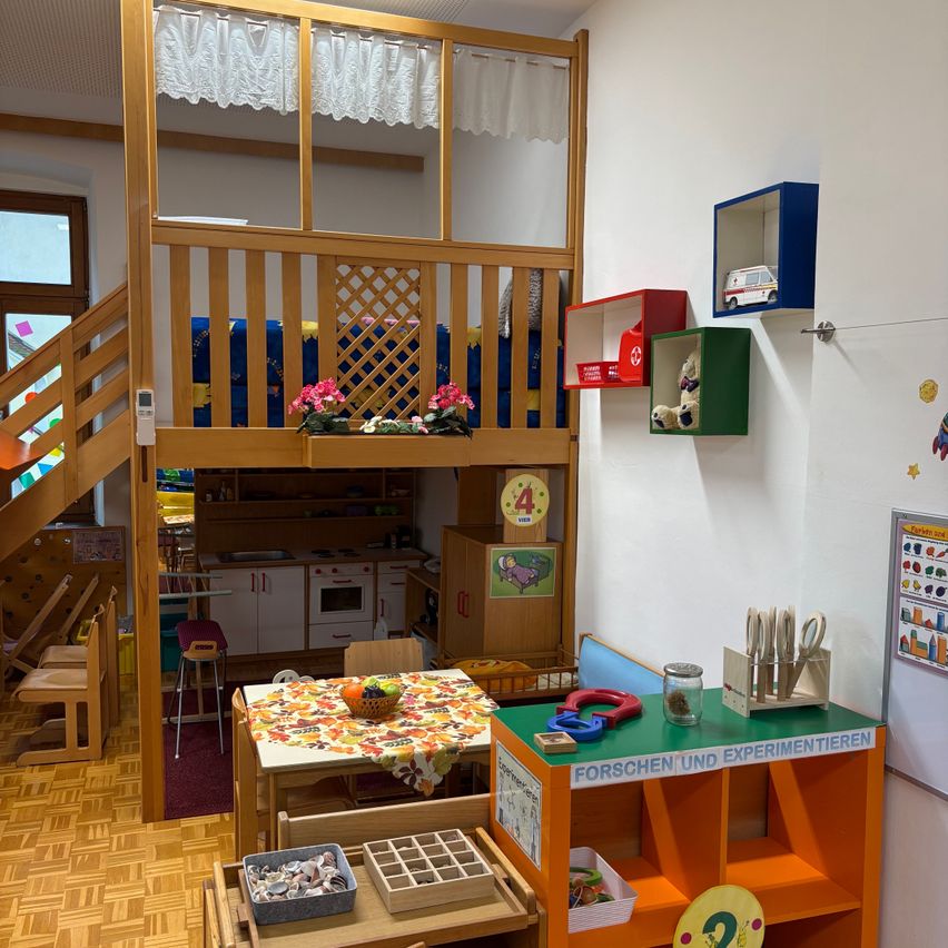 A playroom with a wooden loft bed, a table with fruits, and shelves with toys and books. The room has a wooden floor and white walls.