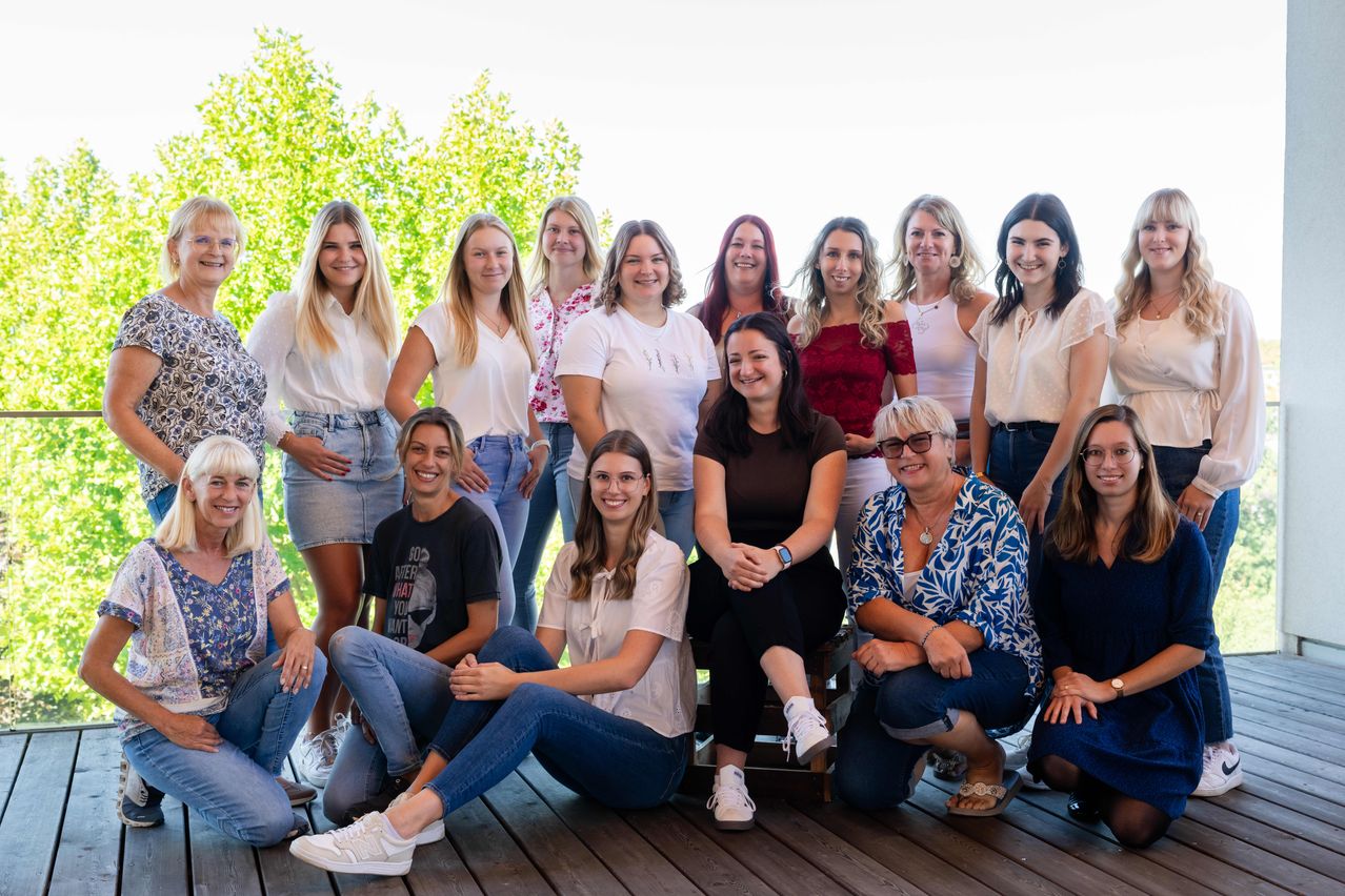 A group of women, some standing and some sitting, pose for a photo on a wooden deck with trees in the background.