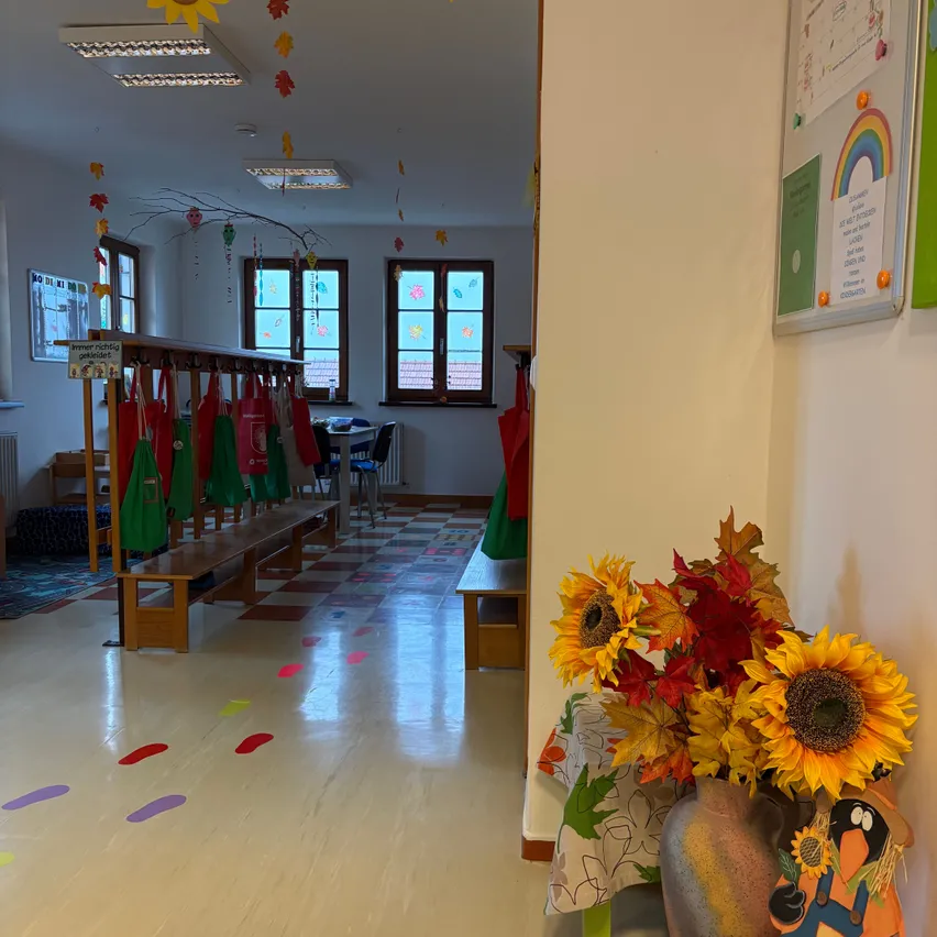 Ein Klassenzimmer mit Holzbänken, bunten Fußabdrücken auf dem Boden und einem Blumenstrauß aus Sonnenblumen in einer Vase auf einem Tisch. Der Raum hat weiße Wände, Fenster und eine Anschlagtafel mit einem Regenbogen und verschiedenen Papieren.