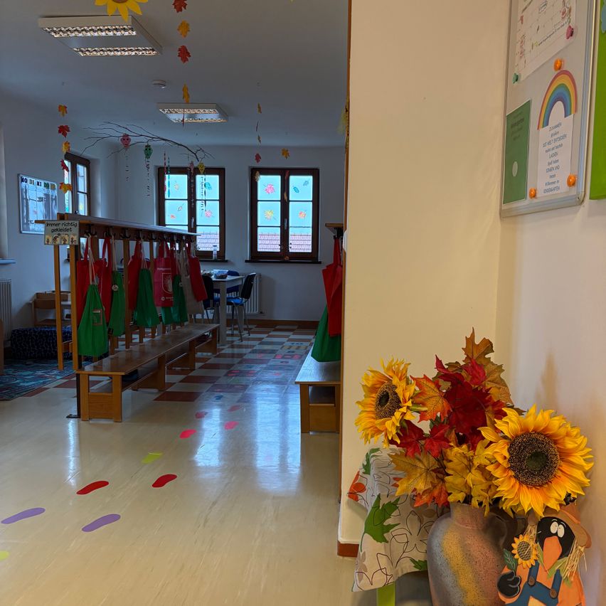 A classroom with wooden benches, colorful footprints on the floor, and a vase of sunflowers on a table. The room has white walls, windows, and a bulletin board with a rainbow and various papers.