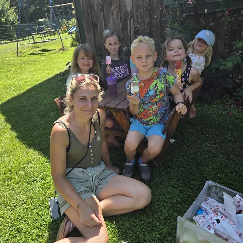Eine Familie posiert für ein Foto im Garten. Eine Frau sitzt auf dem Rasen mit ihren Kindern um einen Tisch herum. Sie halten Eispops in den Händen.